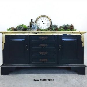 May include: A black painted sideboard with gold accents and five drawers. The sideboard has two doors and sits on four legs. The top of the sideboard is decorated with a garland of greenery and pine cones.