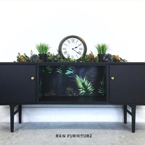 May include: Black mid-century modern sideboard with two doors and a shelf. The shelf is lined with tropical leaf patterned wallpaper. The sideboard has gold knobs and sits on four legs.