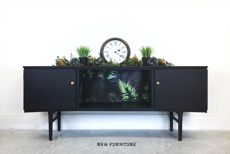 May include: Black mid-century modern sideboard with two doors and a shelf. The shelf is lined with tropical leaf patterned wallpaper. The sideboard has gold knobs and sits on four legs.
