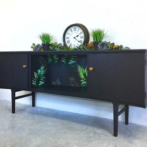 May include: A black mid-century modern sideboard with two doors and a glass-front cabinet. The cabinet has a tropical leaf patterned wallpaper. The sideboard has brass knobs and sits on four legs.