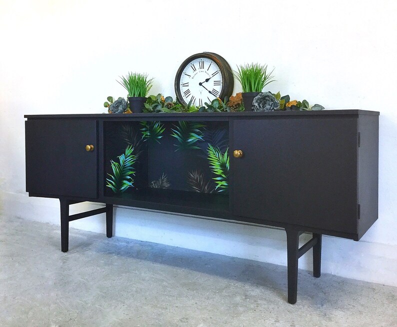 May include: A black mid-century modern sideboard with two doors and a glass-front cabinet. The cabinet has a tropical leaf patterned wallpaper. The sideboard has brass knobs and sits on four legs.
