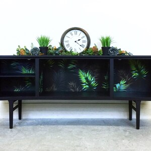 May include: Black sideboard with two doors and a shelf inside. The doors are open and the inside is lined with tropical leaf patterned wallpaper. The sideboard has a clock and plants on top.