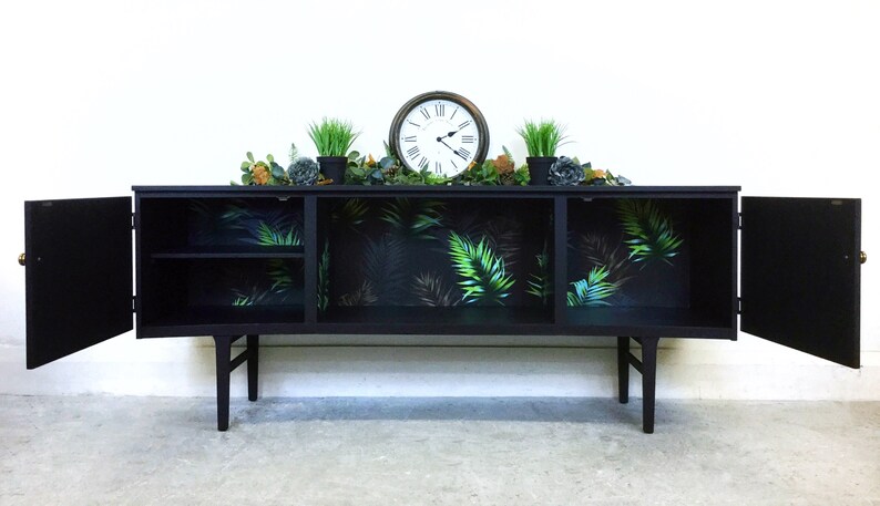 May include: Black sideboard with two doors and a shelf inside. The doors are open and the inside is lined with tropical leaf patterned wallpaper. The sideboard has a clock and plants on top.
