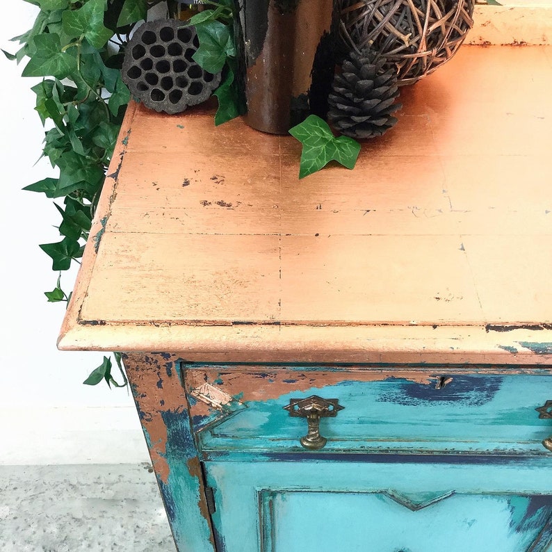 Op de afbeelding: Een vintage kast met een verweerde turquoise blauwe afwerking en een koperen bovenblad. De kast heeft twee deuren en een lade met antieke messing beslag.