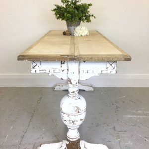 Op de afbeelding: Witte eettafel in landelijke stijl met een verweerde afwerking. De tafel heeft een enkele poot met een gedraaide poot en een rechthoekig blad.