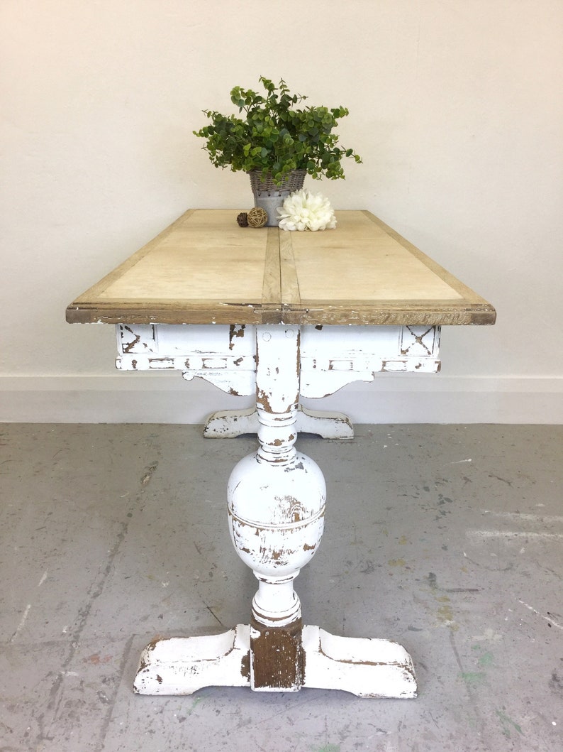 Op de afbeelding: Witte eettafel in landelijke stijl met een verweerde afwerking. De tafel heeft een enkele poot met een gedraaide poot en een rechthoekig blad.