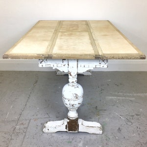 Op de afbeelding: Een wit geschilderde houten tafel met een verweerde afwerking. De tafel heeft een rechthoekig blad met drie bladen die kunnen worden uitgeklapt. De tafel heeft een sokkel met een gedraaide poot en een gesneden ontwerp.