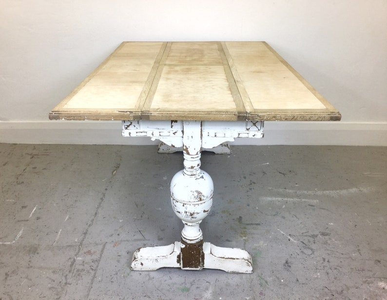 Op de afbeelding: Een wit geschilderde houten tafel met een verweerde afwerking. De tafel heeft een rechthoekig blad met drie bladen die kunnen worden uitgeklapt. De tafel heeft een sokkel met een gedraaide poot en een gesneden ontwerp.