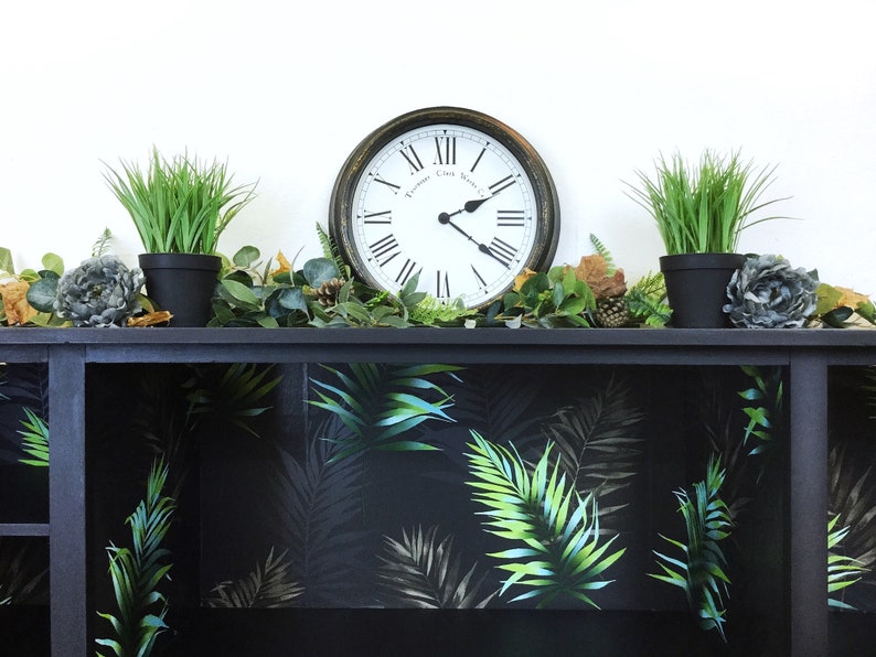 May include: Black shelf unit with a tropical leaf pattern wallpaper lining the shelves. The shelf is decorated with potted plants, a clock, and other decorative items.