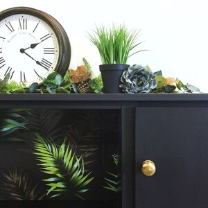 May include: Black cabinet with a single door and a gold knob. The cabinet has a glass front with a tropical leaf pattern wallpaper. The cabinet is decorated with a clock, potted plants, and greenery.