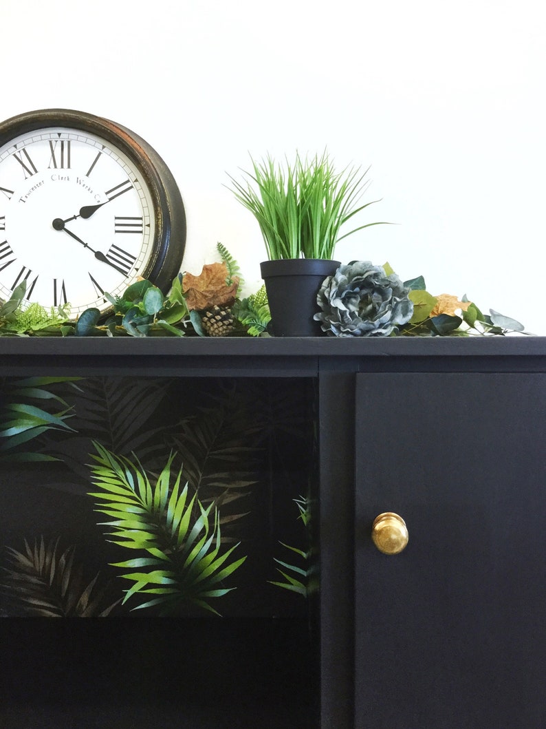 May include: Black cabinet with a single door and a gold knob. The cabinet has a glass front with a tropical leaf pattern wallpaper. The cabinet is decorated with a clock, potted plants, and greenery.