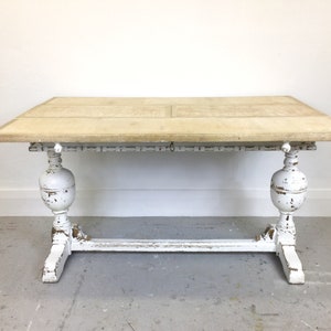 Op de afbeelding: Een wit geschilderde houten tafel met een verweerde afwerking. De tafel heeft een rechthoekig blad en een onderstel met twee gedraaide poten en een dwarsbalk.