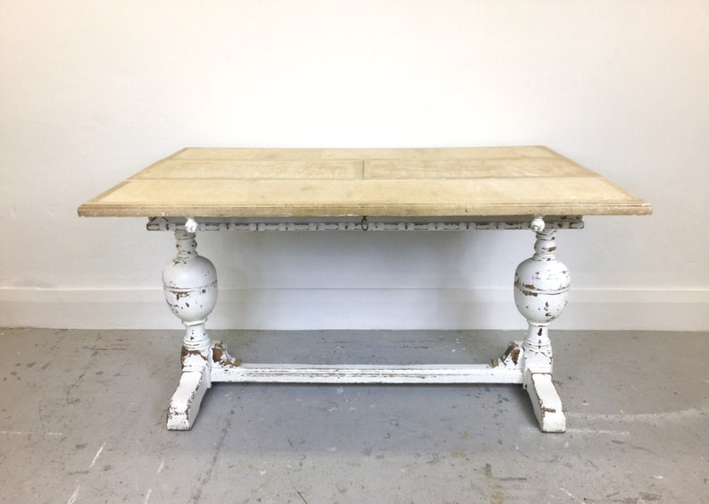 Op de afbeelding: Een wit geschilderde houten tafel met een verweerde afwerking. De tafel heeft een rechthoekig blad en een onderstel met twee gedraaide poten en een dwarsbalk.