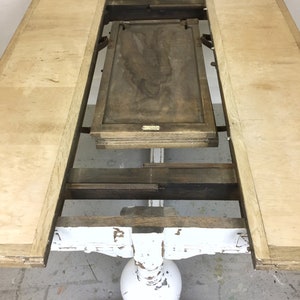Op de afbeelding: Een wit geschilderde houten tafel met een uitklapbare blad. De tafel heeft een verweerde afwerking en een ronde poot. De tafel is open met het blad uitgeklapt, waardoor de onderkant van het tafelblad zichtbaar is.