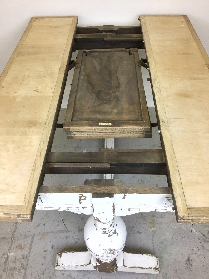 Op de afbeelding: Een wit geschilderde houten tafel met een uitklapbare blad. De tafel heeft een verweerde afwerking en een ronde poot. De tafel is open met het blad uitgeklapt, waardoor de onderkant van het tafelblad zichtbaar is.