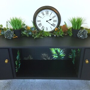 May include: Black cabinet with two doors and a shelf. The shelf is lined with tropical leaf patterned wallpaper. The cabinet has gold knobs. A clock and potted plants are on top of the cabinet.
