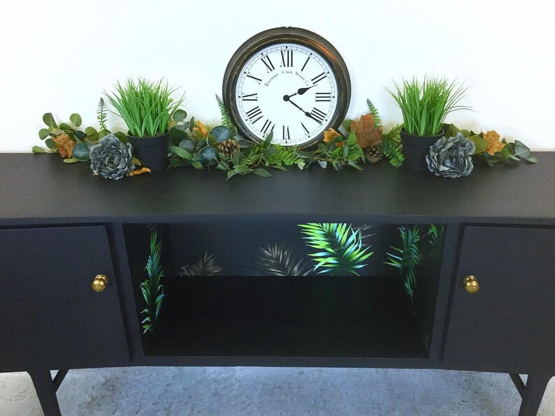 May include: Black cabinet with two doors and a shelf. The shelf is lined with tropical leaf patterned wallpaper. The cabinet has gold knobs. A clock and potted plants are on top of the cabinet.