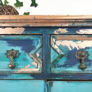 Op de afbeelding: Een verweerde teal en koperen geschilderde commode met twee laden en sierlijke messingknoppen. De commode heeft een verweerde afwerking met afbladderende verf en zichtbare houtnerf.