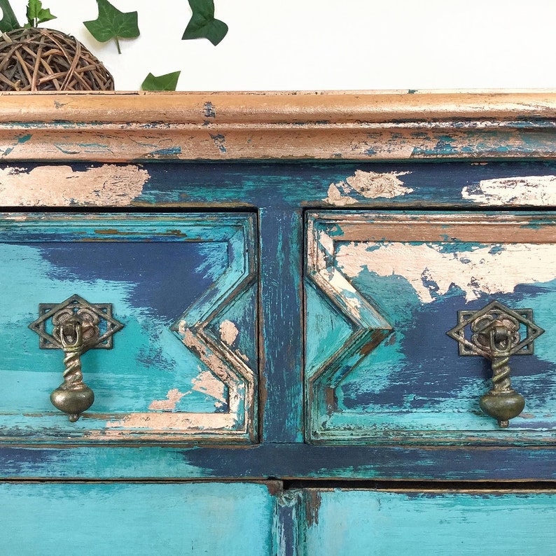 Op de afbeelding: Een verweerde teal en koperen geschilderde commode met twee laden en sierlijke messingknoppen. De commode heeft een verweerde afwerking met afbladderende verf en zichtbare houtnerf.