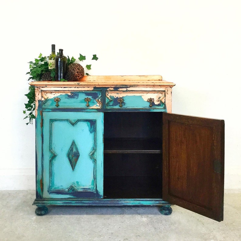 Op de afbeelding: Een verweerde turquoise en koperen kast met twee laden en een deur. De kast heeft een verweerde afwerking en is versierd met een ruitpatroon op de deuren. De kast is open en het interieur is zichtbaar.
