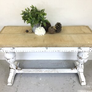 Op de afbeelding: Witte tafel in landelijke stijl met een verweerde afwerking. Het tafelblad is van hout en heeft een lichtbruine vlek. De tafel heeft twee poten met een decoratief ontwerp.