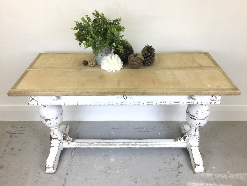 Op de afbeelding: Witte tafel in landelijke stijl met een verweerde afwerking. Het tafelblad is van hout en heeft een lichtbruine vlek. De tafel heeft twee poten met een decoratief ontwerp.