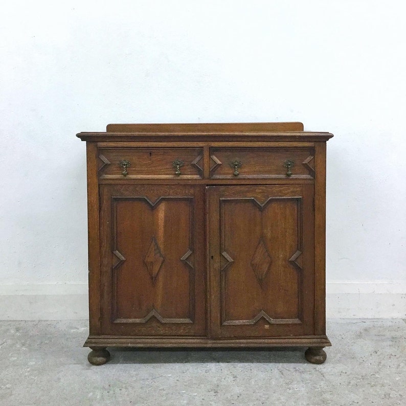 Op de afbeelding: Een vintage houten kast met twee lades en twee deuren. De kast heeft een donkerbruine afwerking en heeft diamantvormige carving op de deuren.