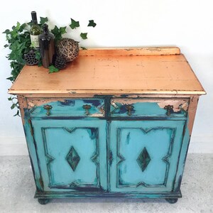 Op de afbeelding: Een verweerd tealblauwe kast met twee laden en twee deuren. De bovenkant van de kast is geschilderd met een koperen afwerking. De kast heeft decoratieve details op de deuren en staat op een witte tegelvloer.