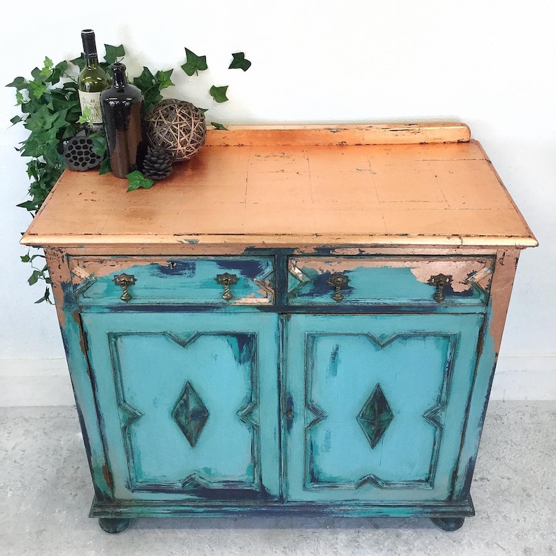 Op de afbeelding: Een verweerd tealblauwe kast met twee laden en twee deuren. De bovenkant van de kast is geschilderd met een koperen afwerking. De kast heeft decoratieve details op de deuren en staat op een witte tegelvloer.