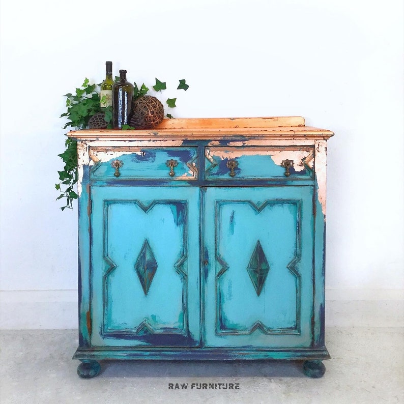Op de afbeelding: Een verweerd turquoise blauwe kast met twee deuren en twee laden. De bovenkant van de kast is koperkleurig geverfd en heeft een verweerde afwerking. De kast staat op een wit oppervlak. RAW FURNITURE is onderaan de afbeelding gedrukt.