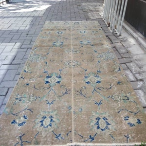 Może przedstawiać: Wzorzysty dywan vintage w kolorze beżowym z kwiatowym wzorem w odcieniach niebieskiego i zielonego. Dywan ma białą frędzlową obwódkę.