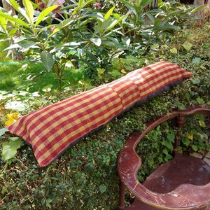 Peut inclure: Un oreiller à carreaux rouges et jaunes avec un dos gris foncé repose sur un lit de feuillage vert. L'oreiller est long et rectangulaire, et il est fait d'un tissu doux et tissé.