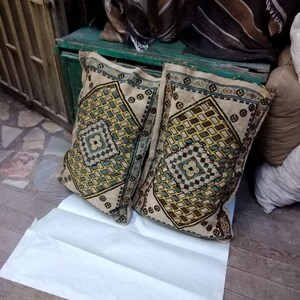 May include: Two decorative pillows with a geometric pattern in shades of brown, beige, green, and blue. The pillows are lying on a white sheet of paper on a wooden floor.