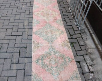 May include: A long, rectangular rug with a faded, vintage appearance. The rug features a pink background with a repeating pattern of green and beige floral medallions. The rug is placed on a brick surface.
