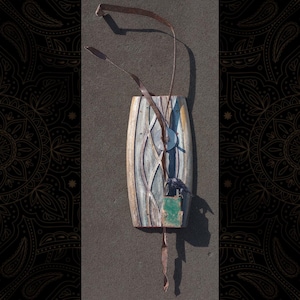 May include: A handcrafted, abstract wall hanging with a weathered, oval-shaped wooden base. It features a geometric design, a small metal disc, and a green square element. Brown leather straps extend from the top and bottom.