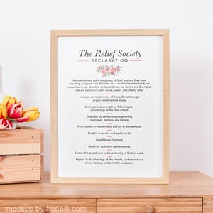 May include: A framed print with the text "The Relief Society DECLARATION" in a decorative font, with floral accents. The text lists values such as faith, virtue, and charity. The frame is light wood, and the print is on a wooden surface.