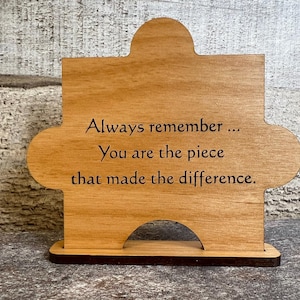 May include: A wooden puzzle piece with the text "Always remember... You are the piece that made the difference." The piece is light brown with a darker brown base. The background is a light gray and white.
