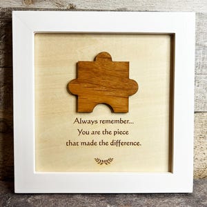 May include: A framed wooden puzzle piece with the text "Always remember... You are the piece that made the difference." The puzzle piece is a warm brown color and is centered on a light wood background within a white frame.
