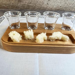 Puede incluir: Una bandeja de madera con cuatro vasos pequeños de chupito de cristal transparente y cuatro trozos de pan blanco. La bandeja es rectangular con una zona empotrada para el pan. Los vasos están vacíos.