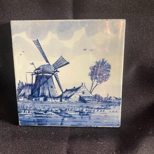 May include: Square ceramic tile featuring a blue and white Dutch windmill scene. The artwork depicts a windmill, houses, and a tree under a cloudy sky. The tile is a decorative piece with a classic, traditional design.