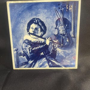 May include: A blue and white ceramic tile featuring a young person playing a flute. A violin and a recorder hang on the wall behind the person. The tile has a square shape and a white border.