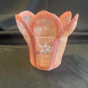 May include: A woven, peach-colored tulip-shaped basket. The basket has a cylindrical base and petal-like edges. A small floral embellishment with white and pink accents decorates the side. The basket is made of a mesh-like material.