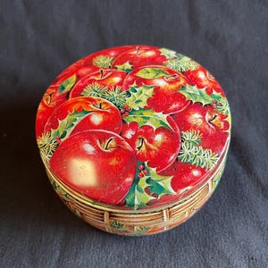 May include: A round, red and green tin with a Christmas-themed design featuring red apples, holly leaves, and pine sprigs. The tin has a gold rim and a woven basket design around the bottom.
