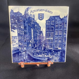 May include: A square ceramic tile featuring a blue and white illustration of Amsterdam's canals and buildings. The word "Amsterdam" is printed at the top, with a coat of arms. A boat is visible on the water.