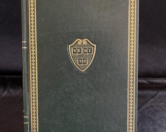 May include: A hardcover book with a dark green cover and gold-colored decorative border. The book features a gold emblem with the text "VERI TAS" on the front cover. The book is propped up on a small stand.