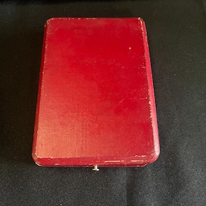 May include: A rectangular, red box with a slightly worn, glossy finish. The box has a small, silver-colored clasp on the front. The box is closed and appears to be empty. The box is sitting on a black surface.