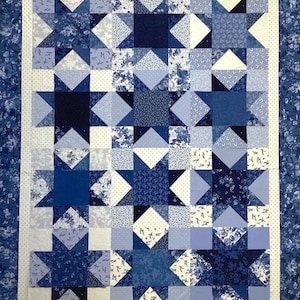 Puede incluir: Un edredón hecho a mano con un diseño de estrellas en varios tonos de azul y blanco. El edredón tiene un borde blanco con pequeños puntos azules y un borde con estampado floral. Los bloques de estrellas están hechos de diferentes telas estampadas.
