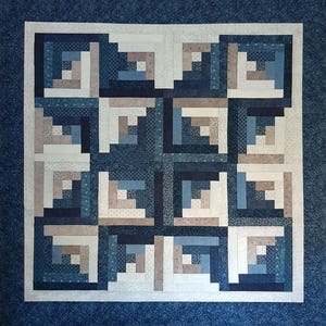 May include: A handcrafted quilt with a log cabin design. The quilt features squares of various shades of blue, white, and tan fabrics. The quilt is bordered by a solid blue fabric.
