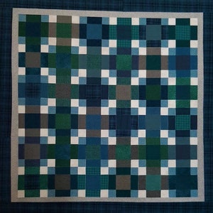 May include: A quilt with a checkered pattern in shades of blue, green, and gray. The quilt is framed with a dark blue border.