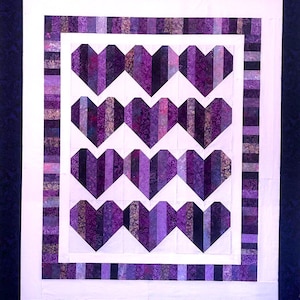 May include: A handmade quilt featuring a repeating heart pattern in shades of purple. The hearts are constructed from vertical strips of fabric, framed by a white border and a striped purple border. The quilt is set against a dark blue background.